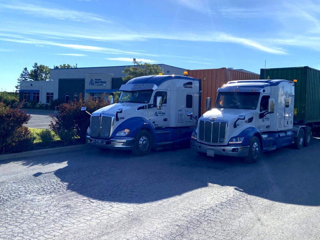 Deux semi-remorques bleus et blancs sont garés côte à côte sur un terrain près d'un bâtiment commercial, chacun transportant un conteneur d'expédition.