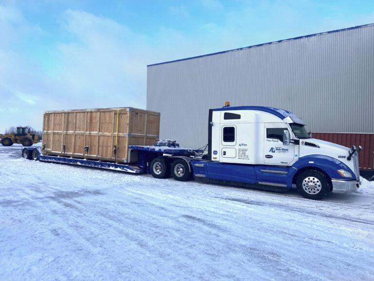Un semi-remorque bleu et blanc avec une remorque à plateau transporte une grande caisse en bois dans une zone industrielle enneigée.