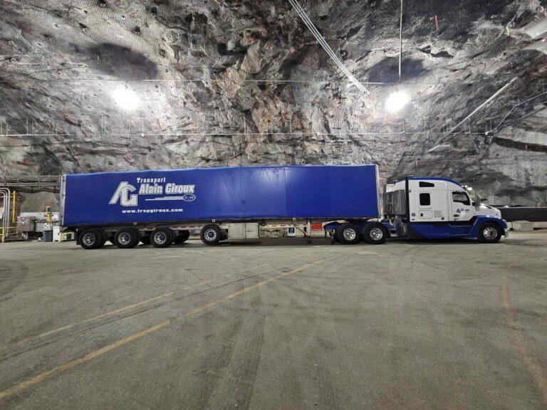 Un gros semi-camion bleu de Transport Alain Giroux est stationné à l'intérieur d'une vaste caverne souterraine aux parois et au plafond rocheux.