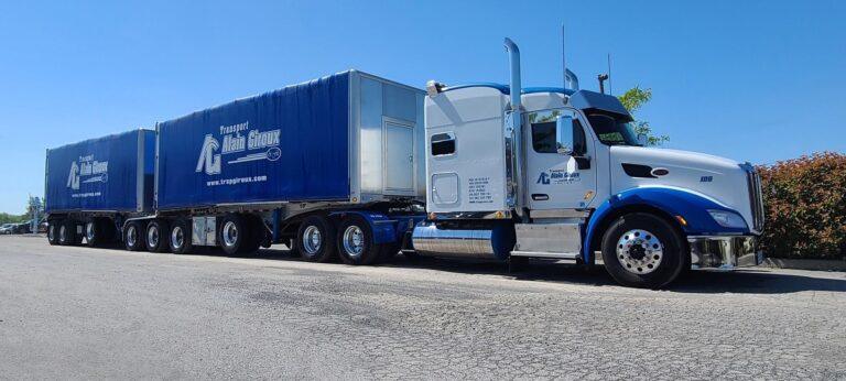 Un semi-camion blanc et bleu avec deux grandes remorques bleues est garé sur une surface asphaltée sous un ciel bleu clair.