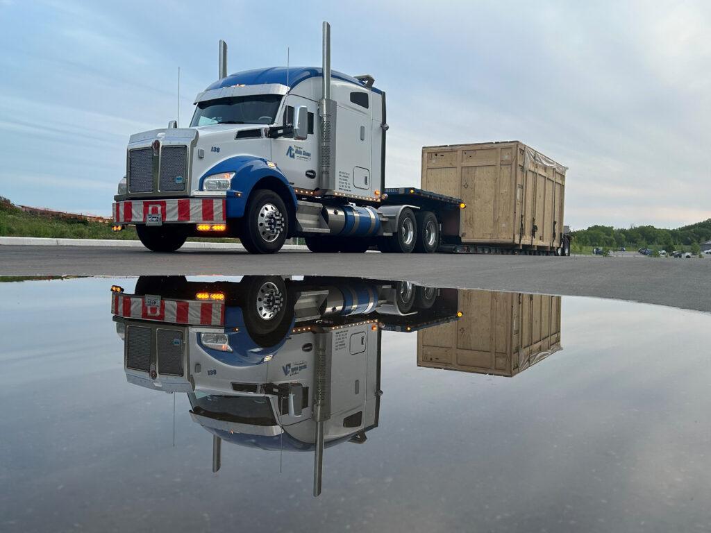 Un semi-remorque bleu et blanc transportant une grande caisse en bois se reflète dans une flaque d'eau par une journée nuageuse.