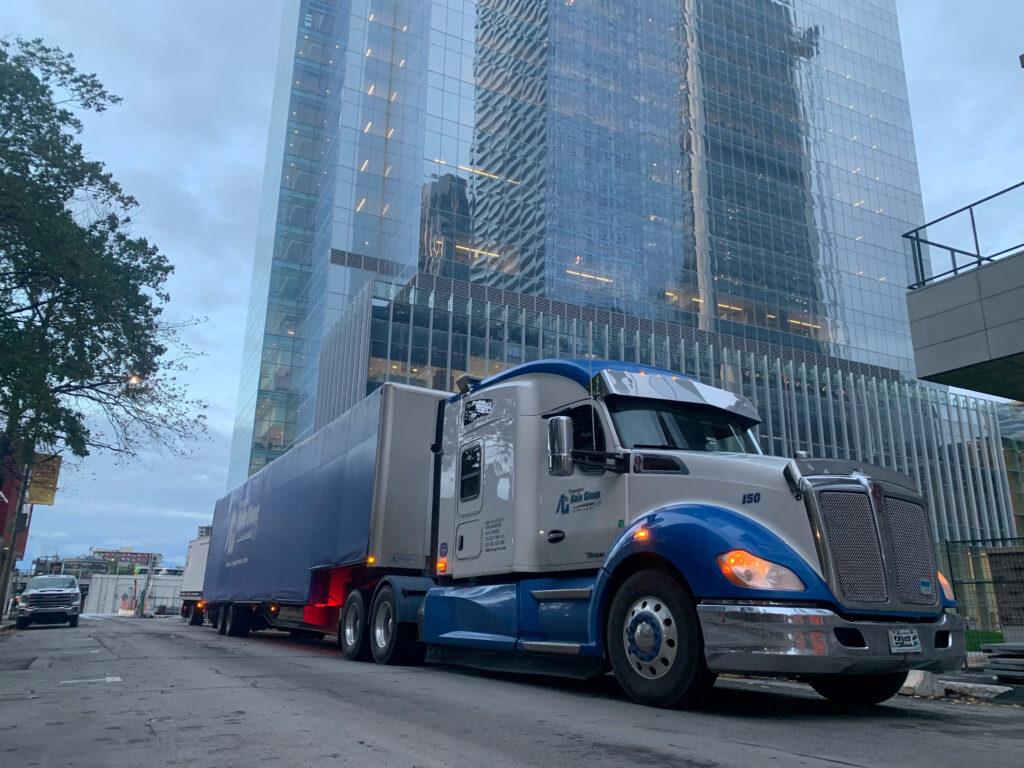 Un gros semi-remorque bleu et blanc avec une remorque est garé dans une rue de la ville devant un grand bâtiment en verre.