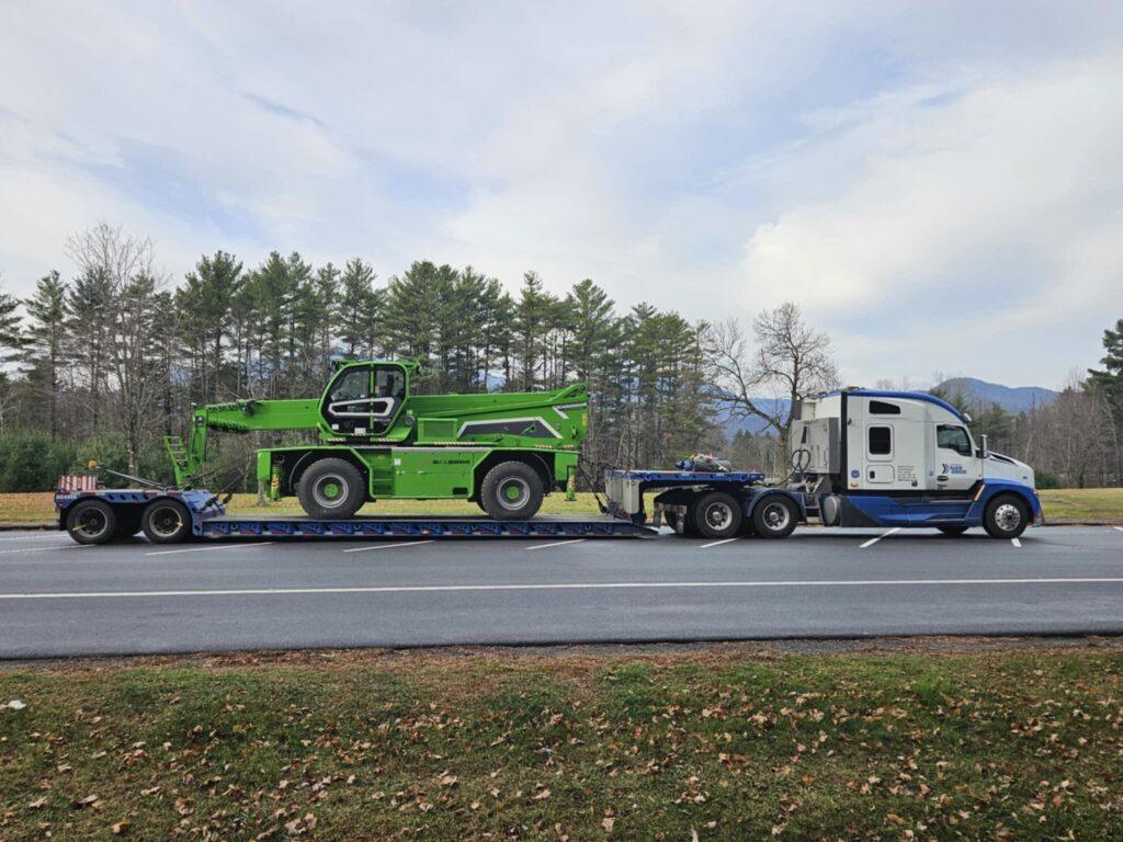 Un semi-remorque blanc transporte un chariot télescopique vert sur une remorque à plateau garée au bord de la route, avec des arbres et des montagnes en arrière-plan.